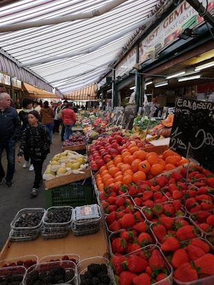 A bustling market scene with a variety of fresh fruits on display. The stall is arranged with vibrant fruits such as strawberries, oranges, and pears, neatly placed in rows. Shoppers are seen walking under a striped canopy roof. Signage with pricing is visible, adding to the lively market atmosphere.