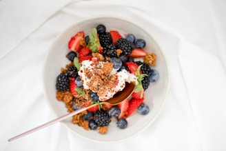 A bowl filled with a mixture of vibrant fresh berries, including strawberries, blueberries, and blackberries, topped with a dollop of yogurt and crunchy granola pieces. A spoon is placed on the side of the bowl, inviting a taste of the colorful and healthy breakfast.
