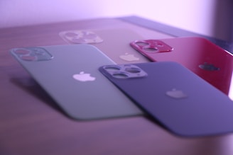 Four smartphone cases with camera cutouts are arranged on a flat surface. The cases are in various colors: green, beige, red, and black, each featuring a logo of a bitten apple. The lighting gives a soft purple hue to the scene.