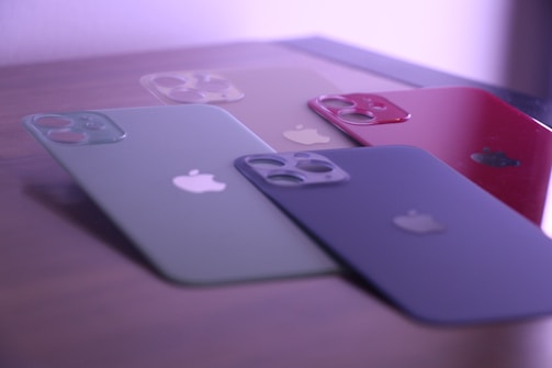 Four smartphone cases with camera cutouts are arranged on a flat surface. The cases are in various colors: green, beige, red, and black, each featuring a logo of a bitten apple. The lighting gives a soft purple hue to the scene.