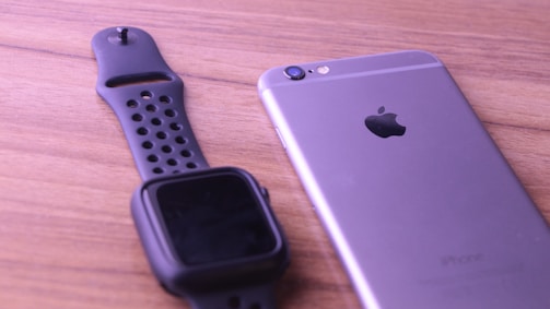 A black smartwatch with a perforated band is placed next to a silver smartphone with a visible Apple logo, all resting on a wooden surface.