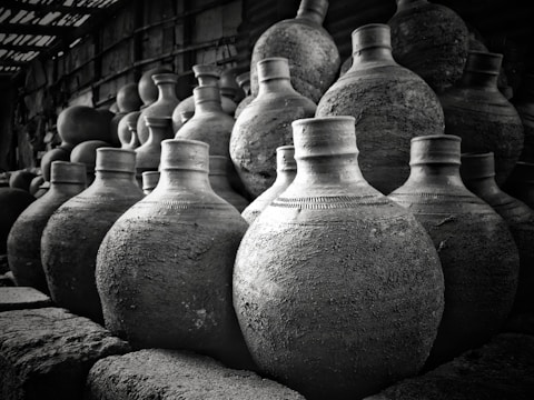A collection of traditional clay pots, stacked closely together, displaying rough textures and varying shades of gray. The lighting creates strong contrasts, highlighting the shape and craftsmanship of each pot.