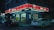 A convenience store situated on a street corner at night. The store has a bright red sign with white text, and several cars are parked in front of it. The store is illuminated by various lights, creating a stark contrast against the dark surroundings. There are posters and advertisements on the store's windows.