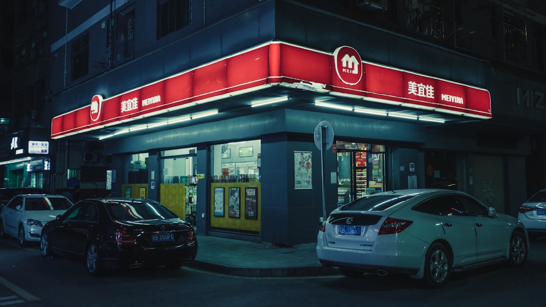 A convenience store situated on a street corner at night. The store has a bright red sign with white text, and several cars are parked in front of it. The store is illuminated by various lights, creating a stark contrast against the dark surroundings. There are posters and advertisements on the store's windows.