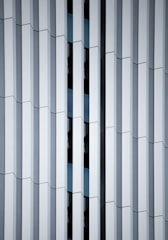 Geometric facade of a building featuring vertical white panels and reflective glass in between, creating a repetitive and structured pattern with varying depths.