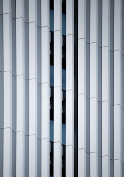 Geometric facade of a building featuring vertical white panels and reflective glass in between, creating a repetitive and structured pattern with varying depths.