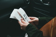 A person holds an open book with both hands, reading intently. The setting appears to be inside a car, indicated by the visible car door and seat. The hands are the primary focus, while the book features bold text and graphic elements.