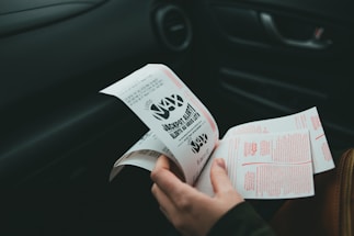 A person is holding and examining what appears to be lottery tickets inside a car. The image is focused on the tickets and the person's hands, with parts of the car interior visible in the background.