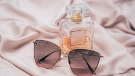 A bottle of Chanel perfume and a pair of stylish sunglasses rest on a soft, silky fabric background. The perfume bottle is square with a golden cap and label, with a subtle peach-colored fragrance inside. The sunglasses have a sleek design with dark, tinted lenses and thin frames.