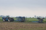 A tractor is parked on a field with a blue agricultural attachment connected to it. The field appears to be flat and stretches into the distance with a few trees along the horizon under an overcast sky.