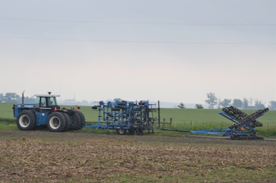 A tractor is parked on a field with a blue agricultural attachment connected to it. The field appears to be flat and stretches into the distance with a few trees along the horizon under an overcast sky.