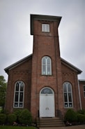 A brick building with a tall, rectangular tower and arched windows on either side of the entrance. The facade features decorative brickwork and a white double door with 'Newport ME Church' above it. Surrounding the building are neatly trimmed bushes and small trees.