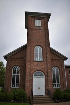 A brick building with a tall, rectangular tower and arched windows on either side of the entrance. The facade features decorative brickwork and a white double door with 'Newport ME Church' above it. Surrounding the building are neatly trimmed bushes and small trees.