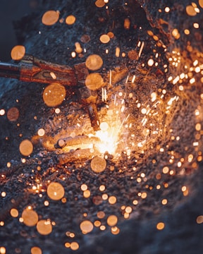 A close-up view of a metal welding process with an industrial torch producing a shower of bright, hot sparks. The image captures the intense light and motion of the welding operation, with vivid orange and yellow sparks contrasting against the dark background.