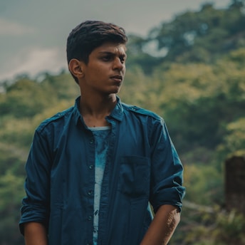 A young person stands outdoors against a backdrop of lush greenery and blurred foliage. They are wearing a blue shirt and appear to be looking off into the distance with a pensive expression.