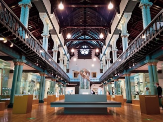 A grand museum interior with tall, ornate columns flanking either side of a central exhibit. The space features a high vaulted ceiling with exposed beams and industrial-style lighting. Glass display cases housing various artifacts are aligned along the walls. A large skeleton exhibit stands prominently at the center, drawing attention. The floors are wooden, and the room has an overall historic and elegant architectural style.