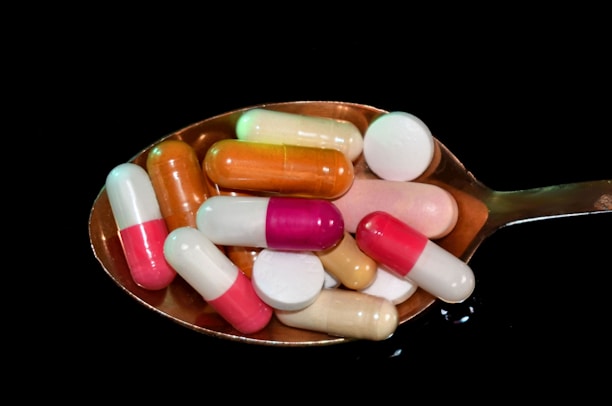 A variety of colorful pills and capsules rest on a metallic spoon against a black background, creating a stark contrast. The pills are of different shapes and sizes, including round tablets and elongated capsules in hues of pink, orange, white, and green.