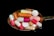 A variety of colorful pills and capsules rest on a metallic spoon against a black background, creating a stark contrast. The pills are of different shapes and sizes, including round tablets and elongated capsules in hues of pink, orange, white, and green.