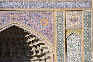 Intricate Islamic architecture featuring ornate geometric patterns and floral motifs in vibrant colors, with a focus on a pointed arch and a vividly decorated facade. The surface includes detailed mosaics and calligraphy.