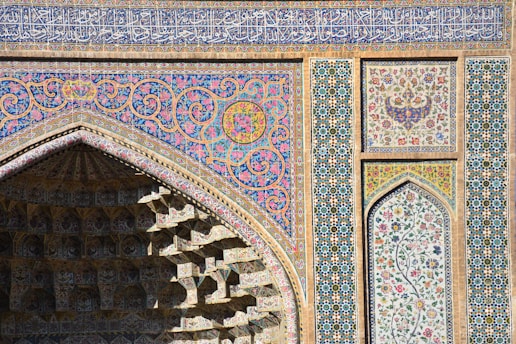Intricate Islamic architecture featuring ornate geometric patterns and floral motifs in vibrant colors, with a focus on a pointed arch and a vividly decorated facade. The surface includes detailed mosaics and calligraphy.
