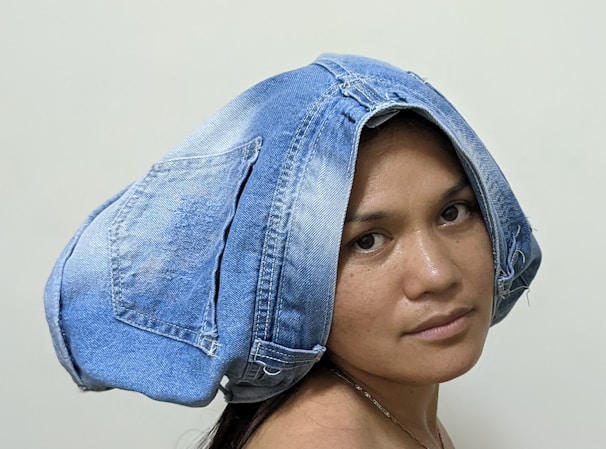A person is wearing a pair of denim jeans on their head as a hat. The jeans appear to be blue with visible stitching and pockets. The background is a plain, light-colored wall.