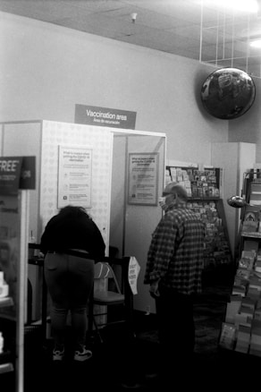 A vaccination area with two people present. One person is standing at a counter reading or signing paperwork, while another stands nearby. This area features informational posters on the walls and appears to be set up in a retail space with greeting card displays visible on the right.