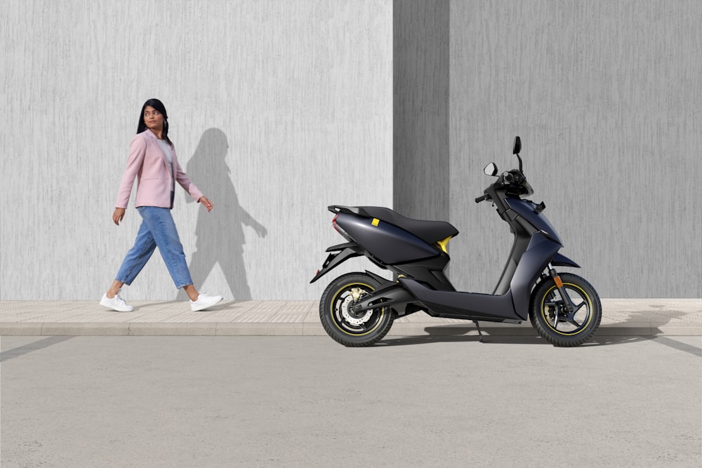 woman in pink jacket and blue denim jeans standing beside black and blue motor scooter