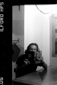 A person with glasses sits at a table reading a book titled 'Mario Puzo Sicilian' in a black and white setting, with subtle wall decorations and a chain in the background.