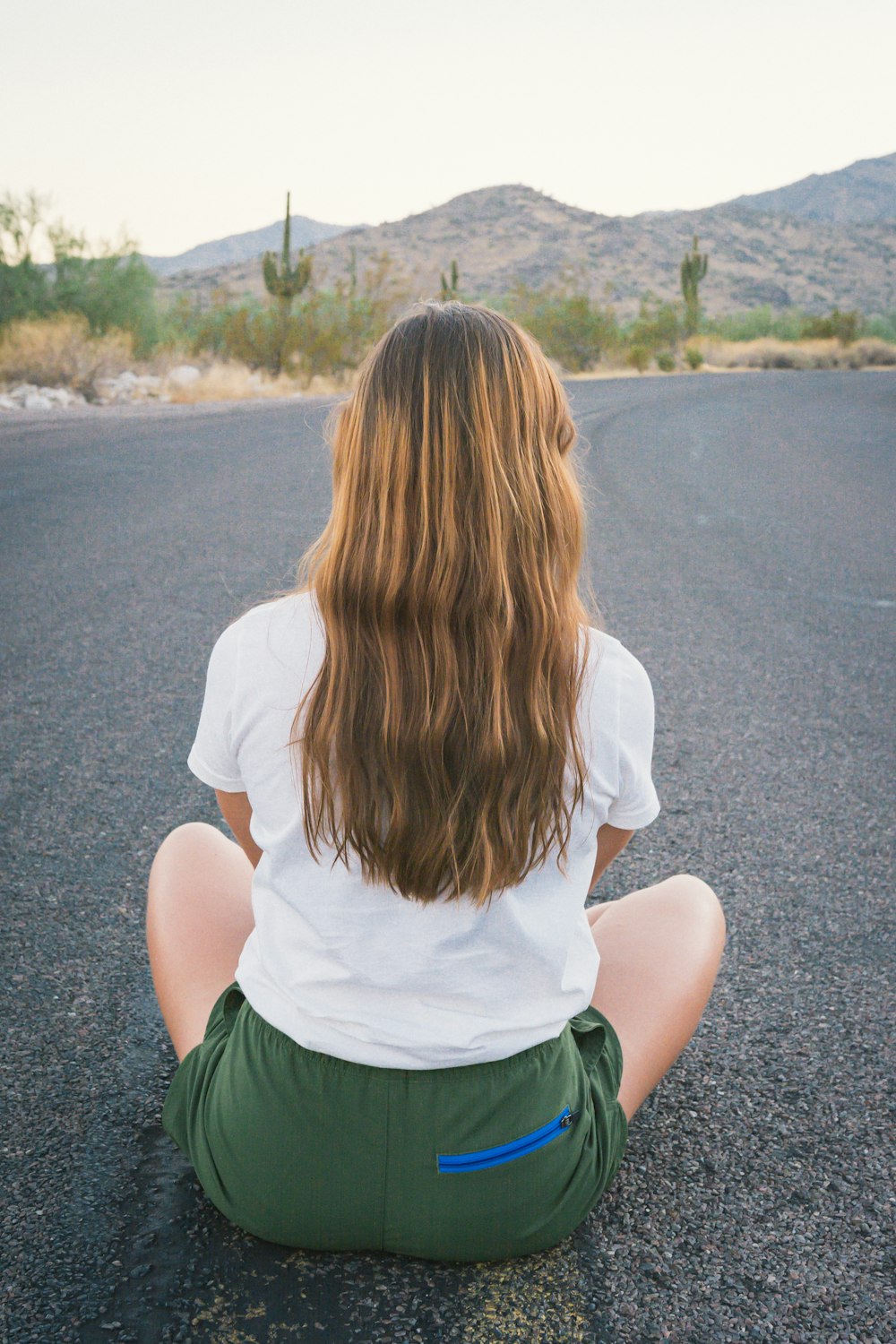 woman in white t-shirt and green shorts sitting on gray concrete road during daytime