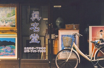 A storefront displays various framed artworks with vivid colors, and a white bicycle with a basket is parked in front. The text on the window is in a non-Latin script, and there are various artistic posters visible through the window. The setting conveys an artistic and quaint atmosphere typical of a small gallery or art shop.