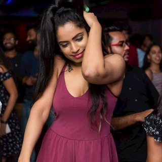 A group of people enjoying themselves in a lively social setting. The central figure is a woman with long dark hair and a burgundy dress, smiling with her head slightly tilted. Others in the background appear to be having fun, contributing to the vibrant atmosphere.