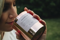 A person is holding a shiny, metallic candle labeled 'NEOPÊCHE' close to their nose, appearing to smell its fragrance. The image has a blurred green background, suggesting an outdoor setting.