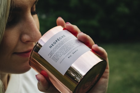 A person is holding a shiny, metallic candle labeled 'NEOPÊCHE' close to their nose, appearing to smell its fragrance. The image has a blurred green background, suggesting an outdoor setting.