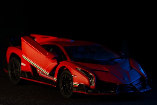 A sleek, red sports car with an aerodynamic design is parked against a dark background. It has sharp, angular features, a low profile, and an open scissor door, showcasing its luxurious and high-performance characteristics. Distinctive stripe detailing runs along its sides, adding to its dynamic appearance.