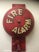 A red fire alarm bell mounted on a wall. The bell is circular with the words 'FIRE ALARM' written in bold, white letters. It has a weathered appearance with some faded and chipped paint.