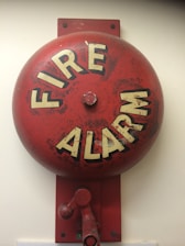 A red fire alarm bell mounted on a wall. The bell is circular with the words 'FIRE ALARM' written in bold, white letters. It has a weathered appearance with some faded and chipped paint.
