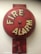 A red fire alarm bell mounted on a wall. The bell is circular with the words 'FIRE ALARM' written in bold, white letters. It has a weathered appearance with some faded and chipped paint.