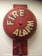 A red fire alarm bell mounted on a wall. The bell is circular with the words 'FIRE ALARM' written in bold, white letters. It has a weathered appearance with some faded and chipped paint.