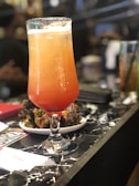 A tall, frosty cocktail glass filled with a gradient orange and red drink is placed on a marble countertop. Beside it, a small white plate holds what appears to be a portion of spicy glazed chicken wings. The background is softly blurred, hinting at a busy bar or restaurant setting.