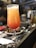 A tall, frosty cocktail glass filled with a gradient orange and red drink is placed on a marble countertop. Beside it, a small white plate holds what appears to be a portion of spicy glazed chicken wings. The background is softly blurred, hinting at a busy bar or restaurant setting.