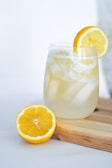 A refreshing drink in a glass with ice cubes, garnished with a lemon slice. Another half lemon lies on a wooden surface beside the glass, creating a bright and inviting scene.