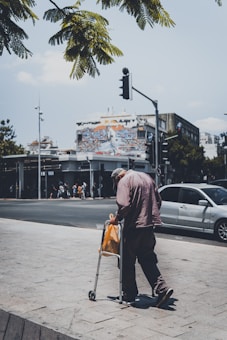 An elderly person using a walker stands by the roadside, holding an orange plastic bag. The scene captures a city intersection with a vibrant mural on a building fa&ccedil;ade in the background. Cars are parked along the street, and a traffic light can be seen. Lush green leaves from a tree partially frame the top of the image.