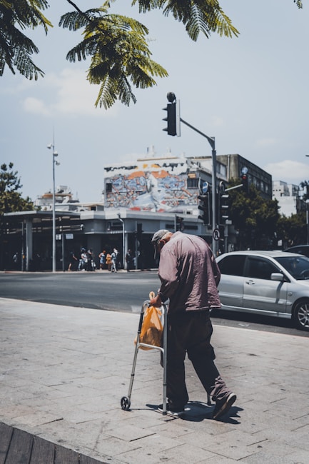 An elderly person using a walker stands by the roadside, holding an orange plastic bag. The scene captures a city intersection with a vibrant mural on a building fa&ccedil;ade in the background. Cars are parked along the street, and a traffic light can be seen. Lush green leaves from a tree partially frame the top of the image.