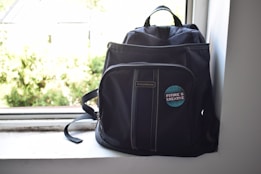 A dark-colored backpack is positioned on a windowsill. The backpack features a badge with the phrase 'Future is Creative' on the front pocket. Sunlight streams in from the window, creating a bright and airy atmosphere.