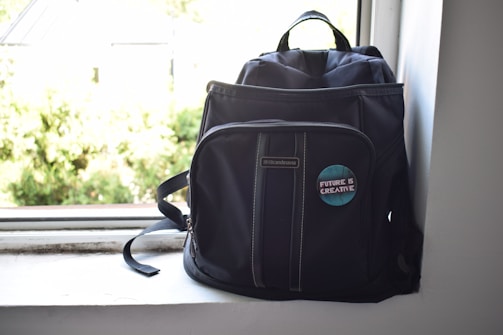 A dark-colored backpack is positioned on a windowsill. The backpack features a badge with the phrase 'Future is Creative' on the front pocket. Sunlight streams in from the window, creating a bright and airy atmosphere.
