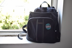 A dark-colored backpack is positioned on a windowsill. The backpack features a badge with the phrase 'Future is Creative' on the front pocket. Sunlight streams in from the window, creating a bright and airy atmosphere.