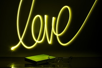 Neon light spelling out the word 'love' in bright yellow against a dark background. Below it, a closed book and a pair of eyeglasses are placed on a reflective surface, casting subtle reflections.
