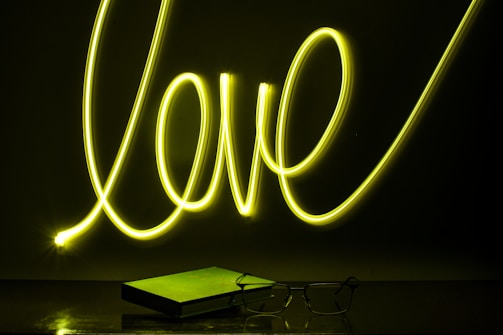 Neon light spelling out the word 'love' in bright yellow against a dark background. Below it, a closed book and a pair of eyeglasses are placed on a reflective surface, casting subtle reflections.