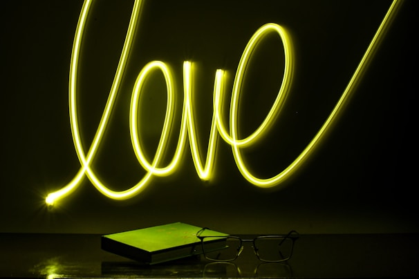 Neon light spelling out the word 'love' in bright yellow against a dark background. Below it, a closed book and a pair of eyeglasses are placed on a reflective surface, casting subtle reflections.