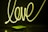 Neon light spelling out the word 'love' in bright yellow against a dark background. Below it, a closed book and a pair of eyeglasses are placed on a reflective surface, casting subtle reflections.